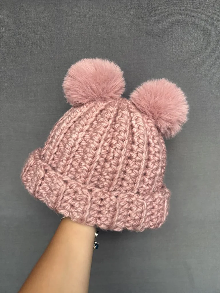 A hand holds a pink crochet hat with two fluffy pom-poms on top, alongside a matching crochet headband, set against a plain gray background.