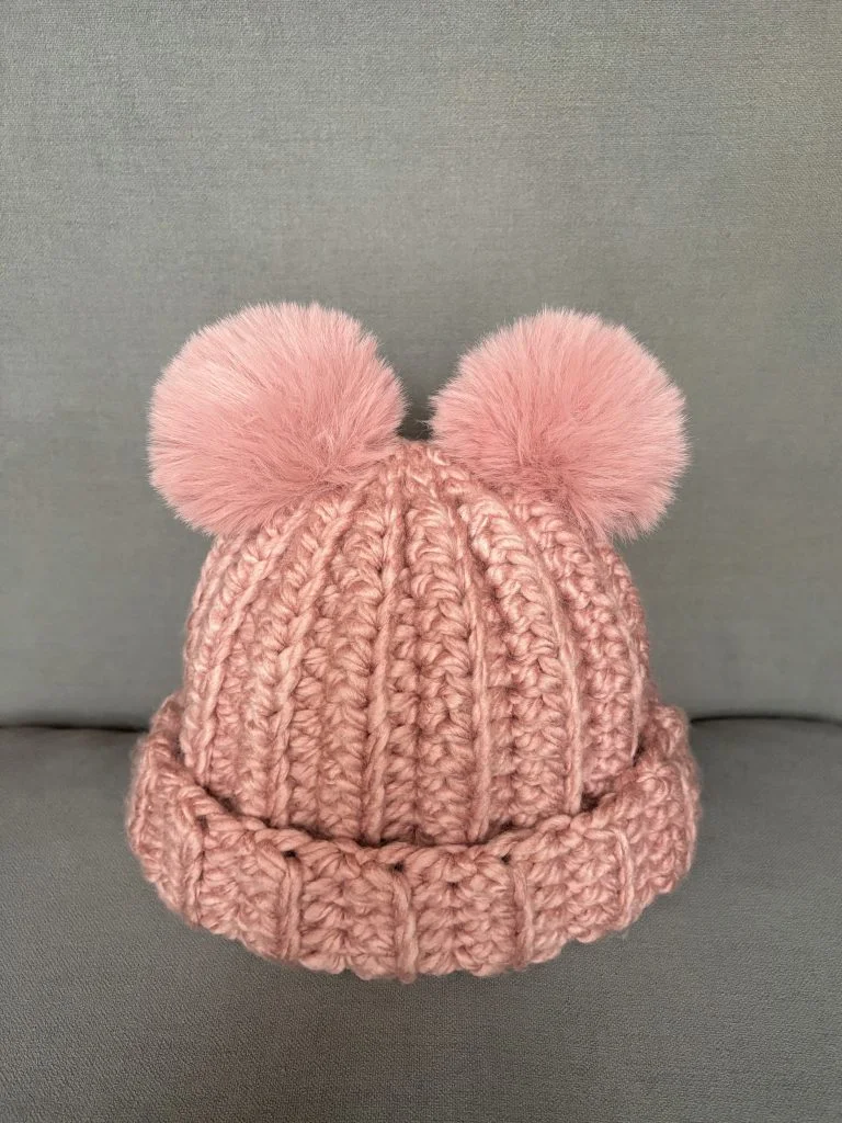 A pink crochet hat with two large fluffy pom-poms on top is displayed against a plain gray background, pairing perfectly with a cozy crochet headband for added warmth and charm.