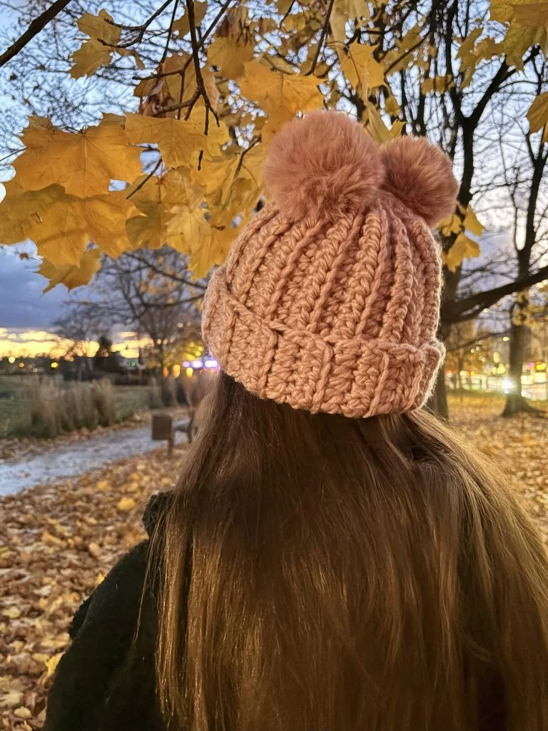A person with long brown hair wearing a pink crochet headband stands outdoors under yellow autumn leaves at sunset.