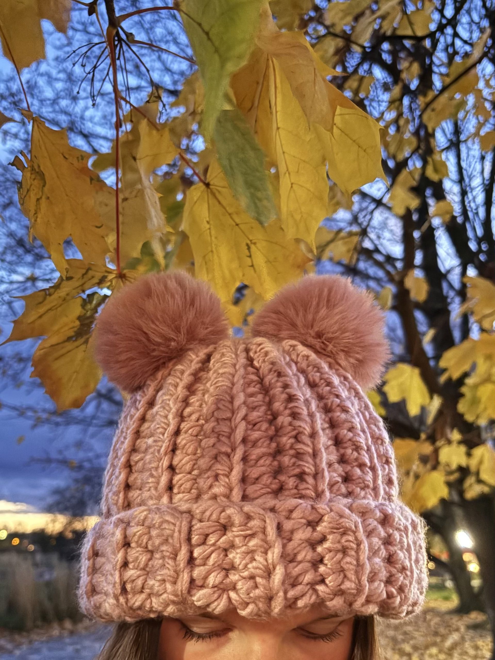 Close-up of a person wearing a chunky pink crochet hat with two pom-poms and a crochet headband, standing outdoors under yellow autumn leaves.