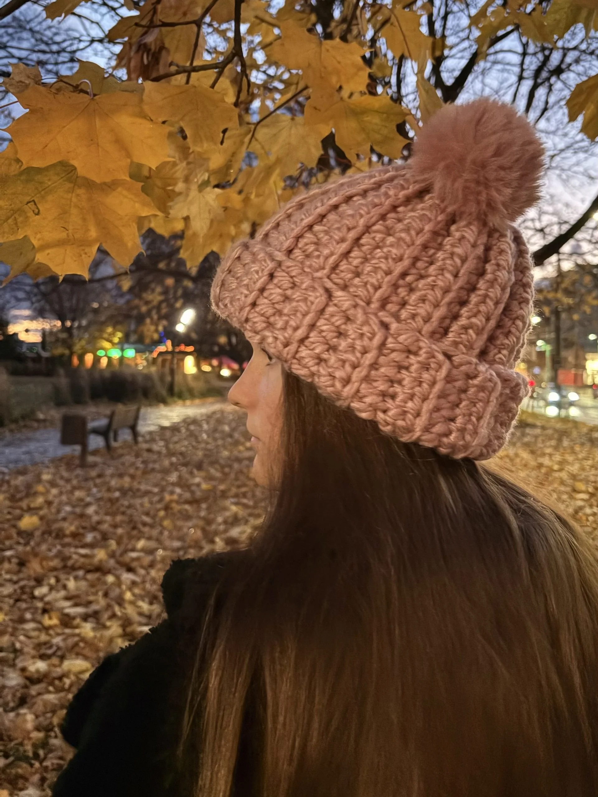 A person with long brown hair wearing a pink crochet headband stands outdoors under autumn leaves near a park path in the evening.