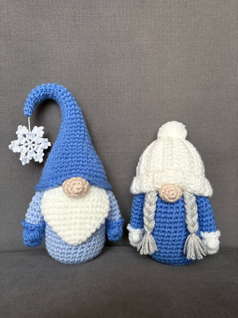 Two crocheted gnome dolls sit side by side; one wears a blue hat with a snowflake and long beard, while the other has a white hat with braids and a simple blue outfit—perfect companions for your next crochet gnome or free crochet pumpkin pattern project.