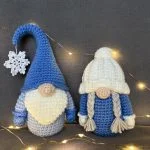 Two crochet gnomes—a tall blue hat with a snowflake and another with white braids—are displayed with string lights on a gray background, perfect for pairing with your favorite pumpkin crochet pattern this season.