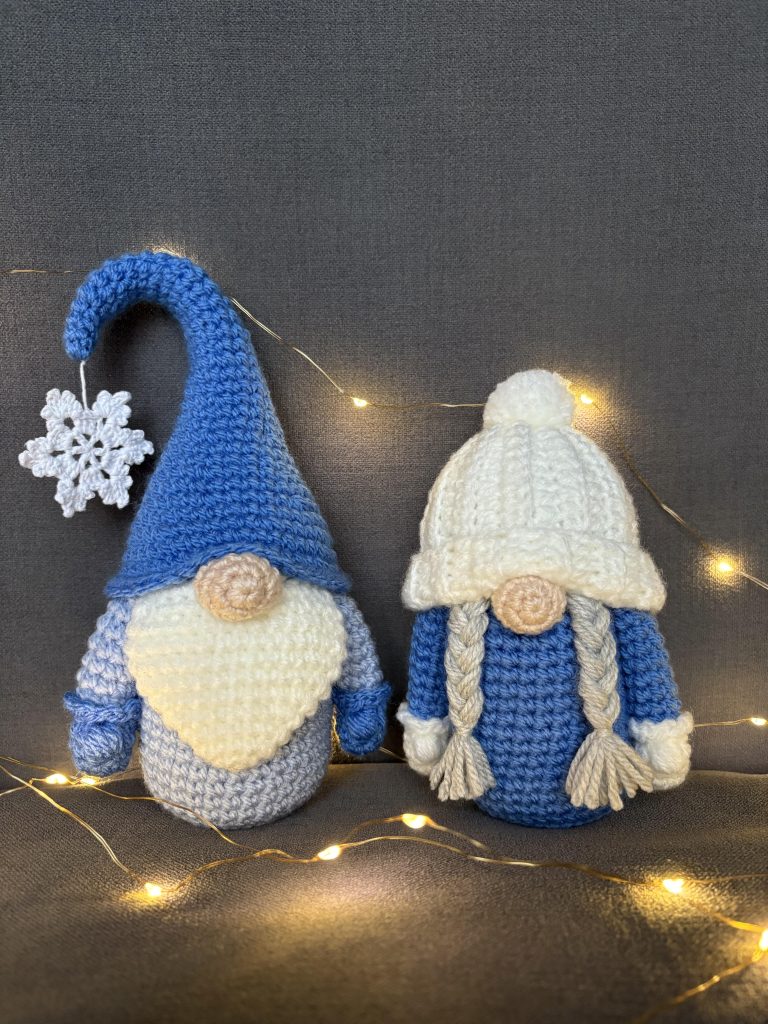 Two crochet gnomes—a tall blue hat with a snowflake and another with white braids—are displayed with string lights on a gray background, perfect for pairing with your favorite pumpkin crochet pattern this season.