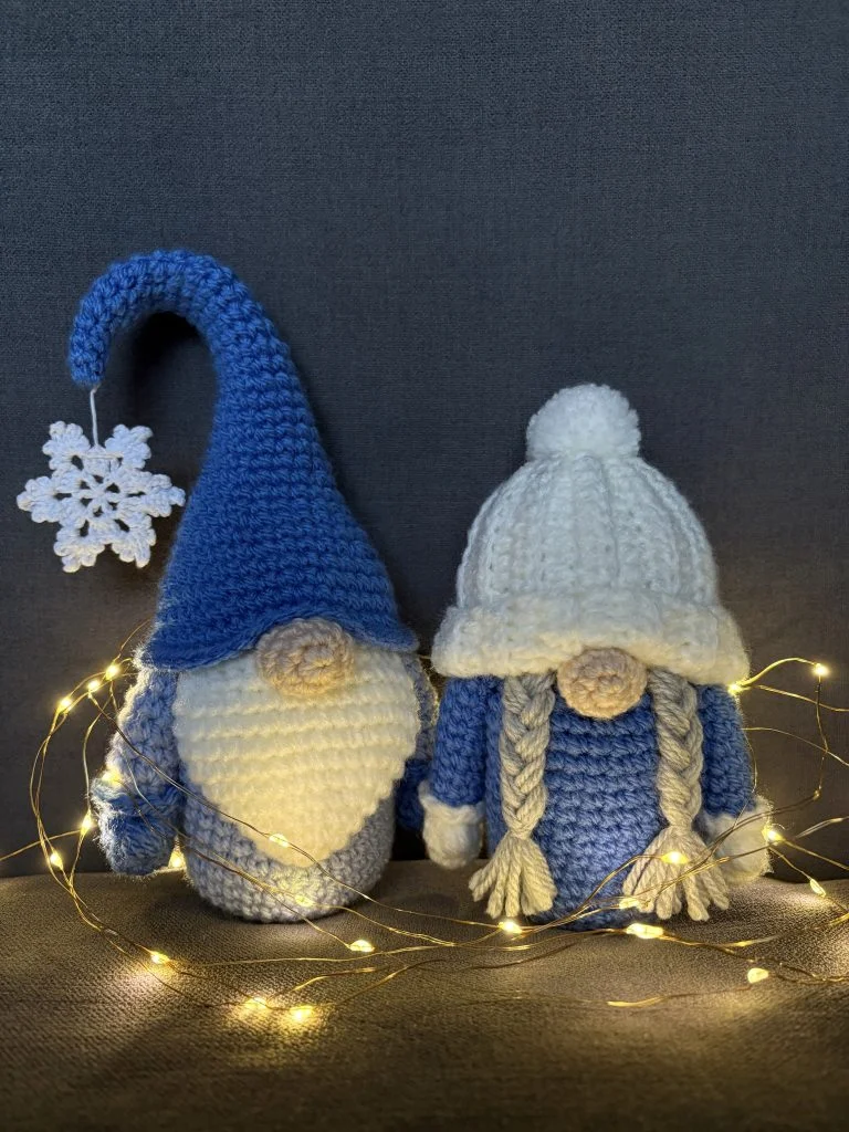 Two crocheted gnome dolls in blue and white with knit hats, surrounded by warm fairy lights, sit on a dark fabric background—perfect inspiration for your next crochet gnome or free crochet pumpkin pattern project.