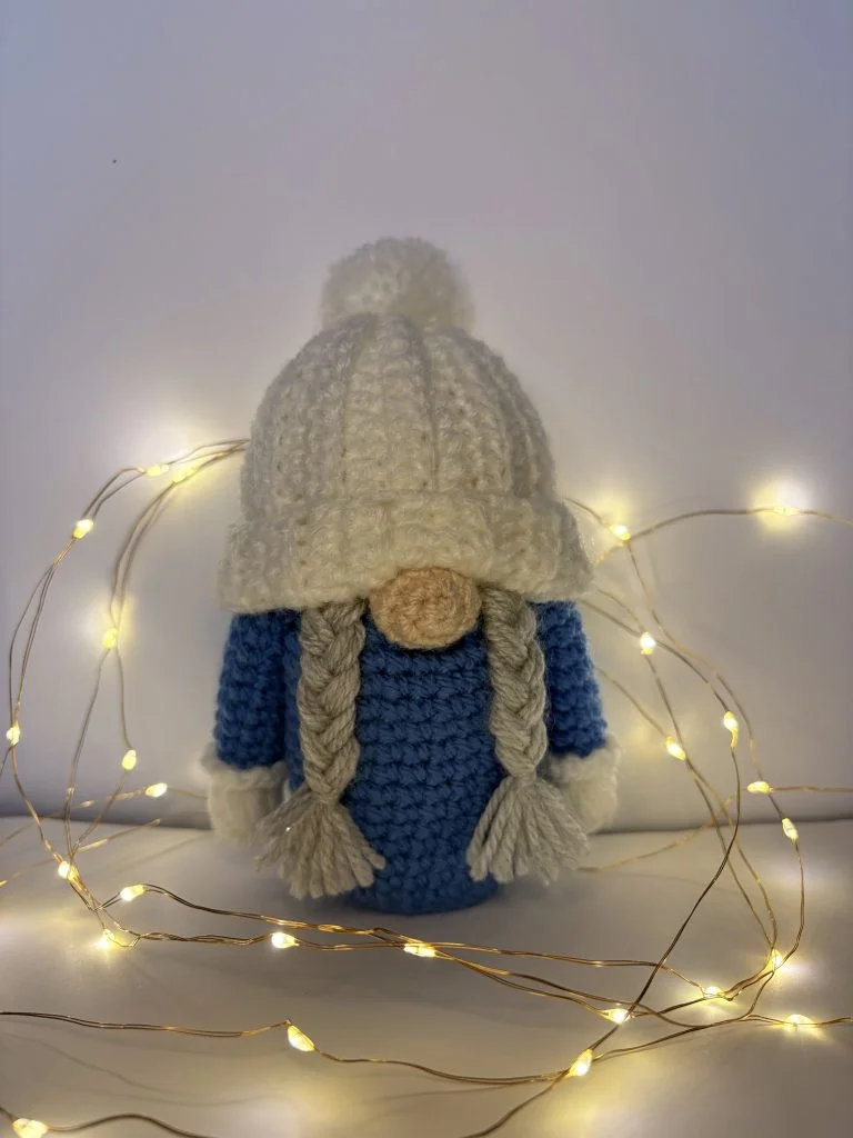 A crocheted doll with long braids, a blue outfit, and a white hat stands surrounded by warm string lights—perfect alongside a crochet gnome or when searching for a free crochet pumpkin pattern to add handmade charm.