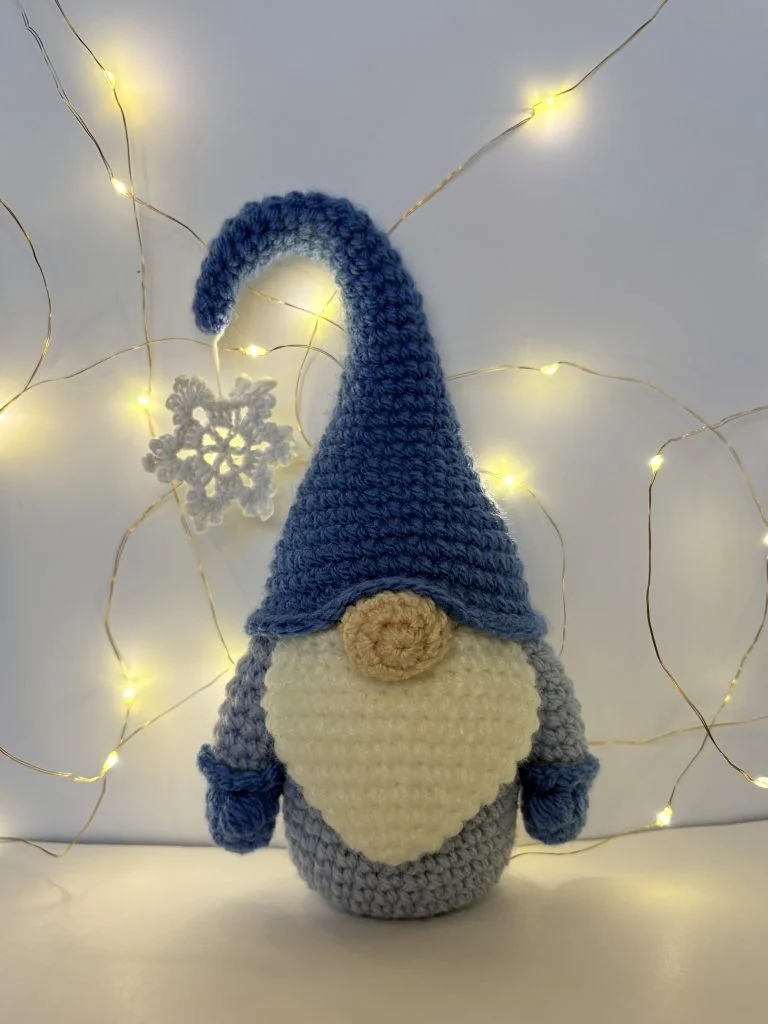 A crochet gnome with a blue hat and white beard stands in front of a white background decorated with string lights and a crocheted snowflake, bringing whimsical charm—explore our crochet pumpkin pattern free for more festive ideas.