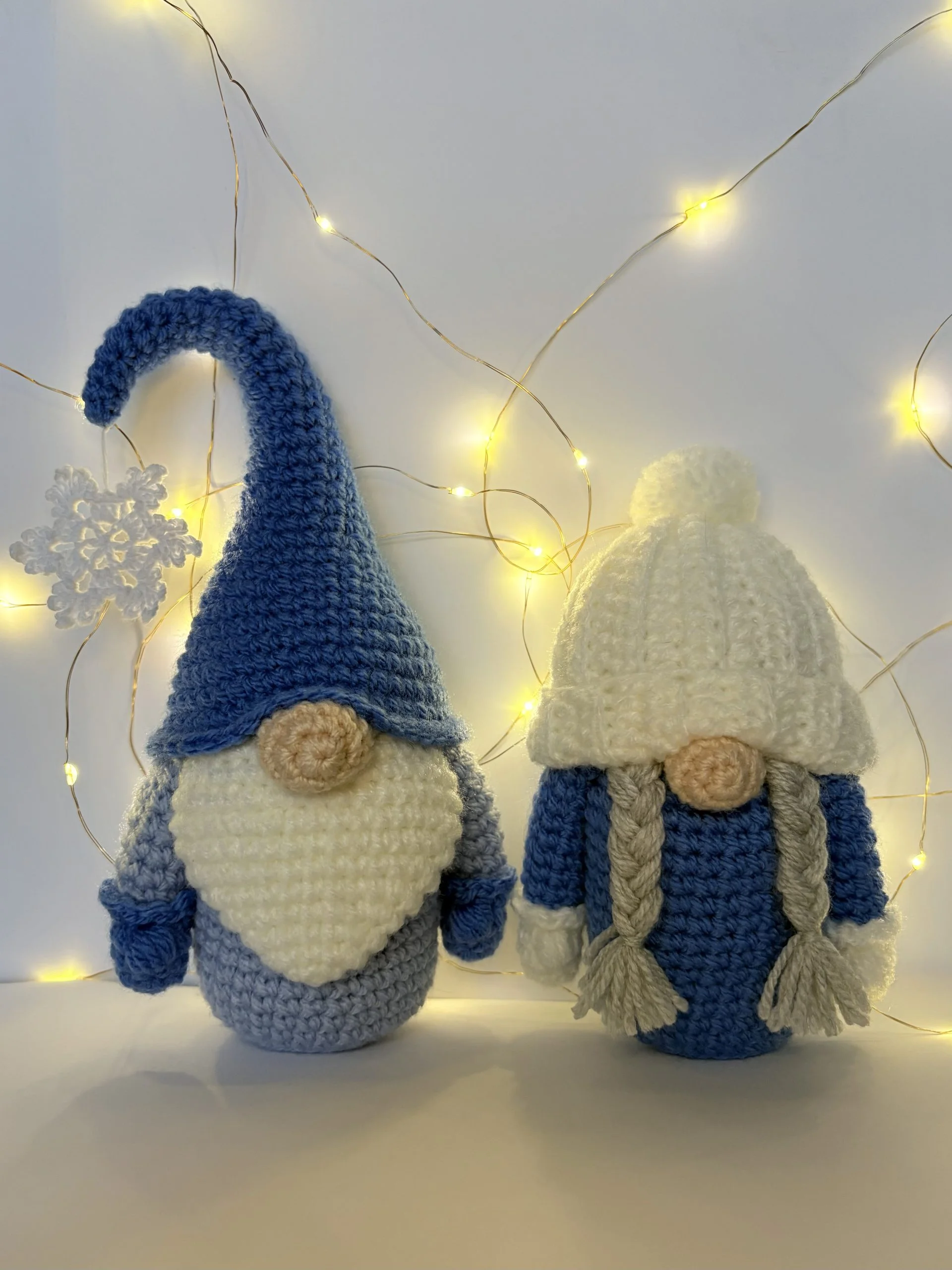 Two crocheted gnome figures, one with a tall blue hat and white beard, the other with a white hat and braids, stand in front of glowing string lights—a cozy scene perfect for showcasing your favorite free crochet pumpkin pattern.