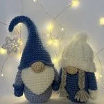 Two crochet gnome dolls with round noses—one in a tall blue hat, the other in a white knit hat with braids—stand before string lights and a snowflake, perfect companions for your next free crochet pumpkin pattern project.