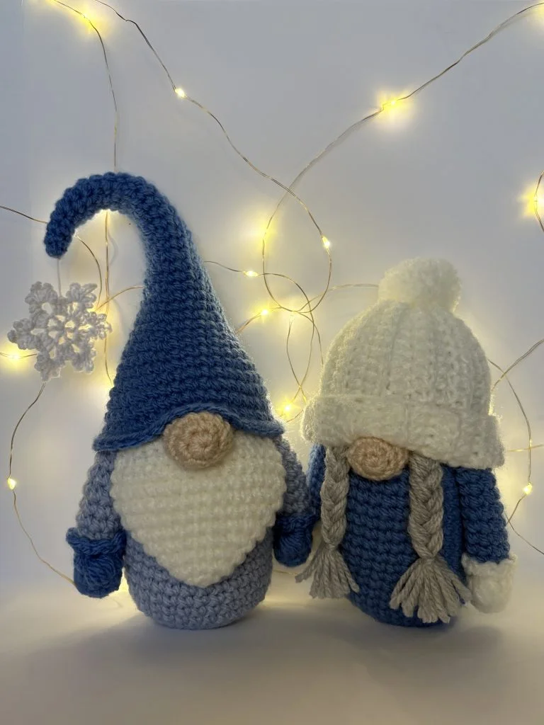 Two crochet gnome dolls with round noses—one in a tall blue hat, the other in a white knit hat with braids—stand before string lights and a snowflake, perfect companions for your next free crochet pumpkin pattern project.