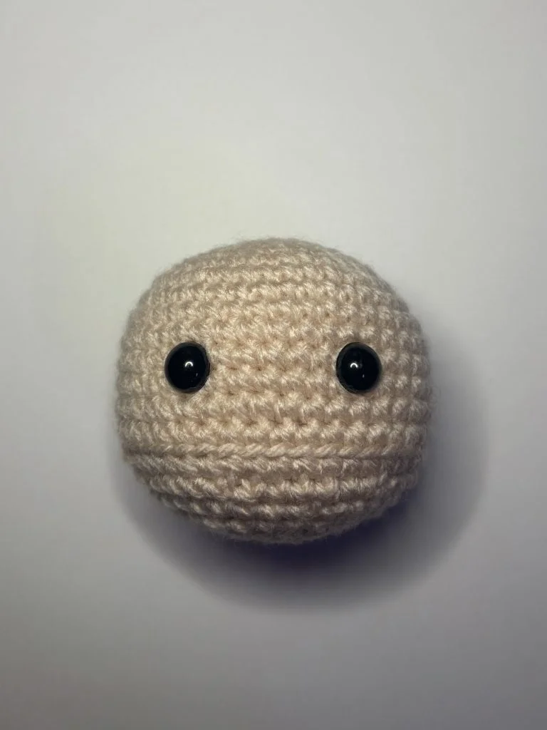 A beige crocheted ball with two black plastic eyes, inspired by a free crochet pumpkin pattern, is placed on a plain white background.