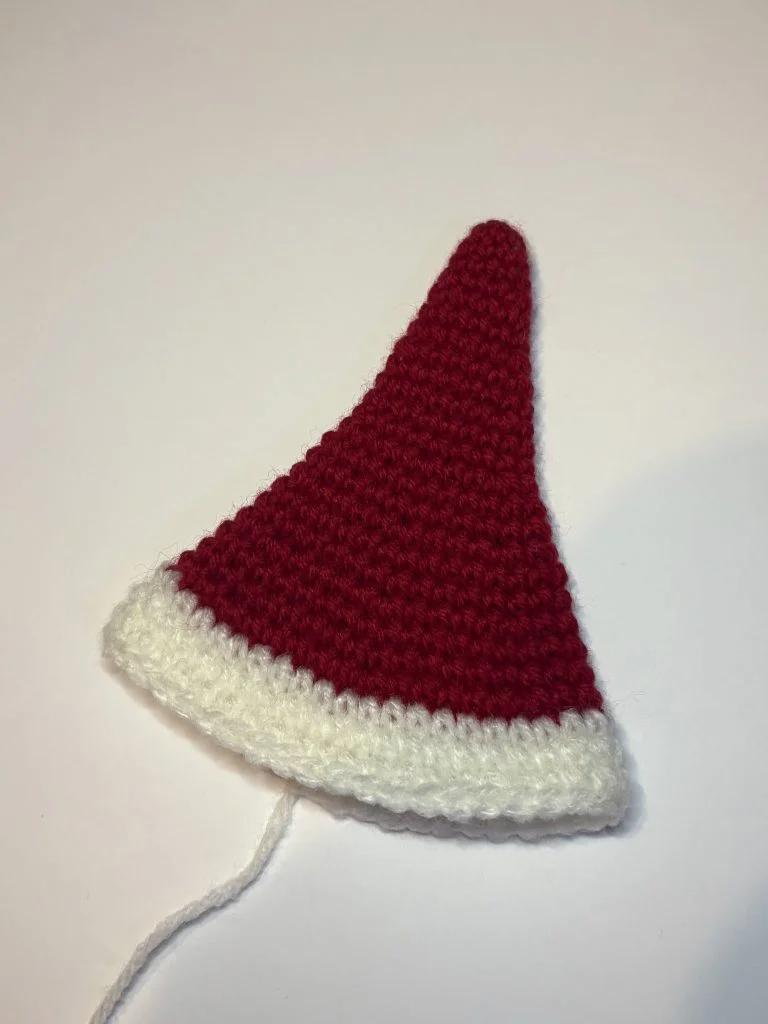 A crocheted red triangle with a white border, resembling the shape of a Santa hat, lies on a plain white surface—an ideal project for those who also enjoy making seasonal items like a crochet pumpkin.