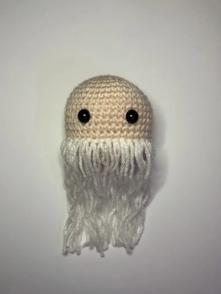 A beige crocheted object with black button eyes and long white yarn strands hanging down, resembling a face with a beard—created using a crochet pumpkin pattern—against a plain white background.