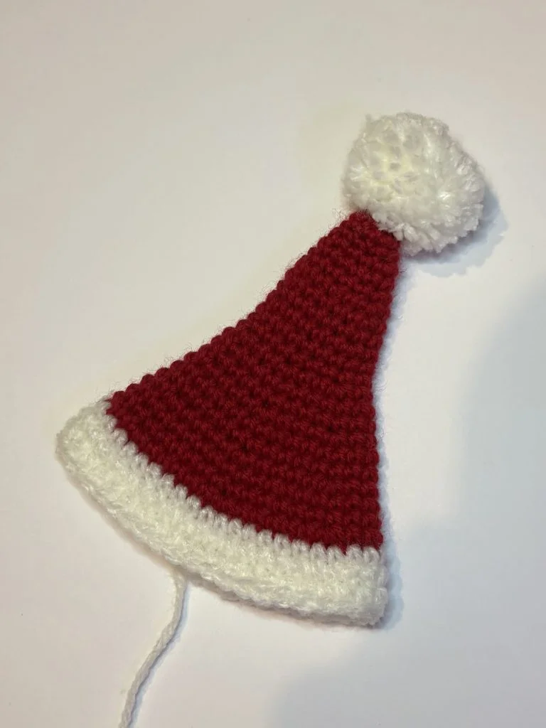 A crocheted Santa hat with a red body, white trim, and a white pom-pom lies on a plain white surface—perfect for pairing with your favorite pumpkin crochet pattern this festive season.