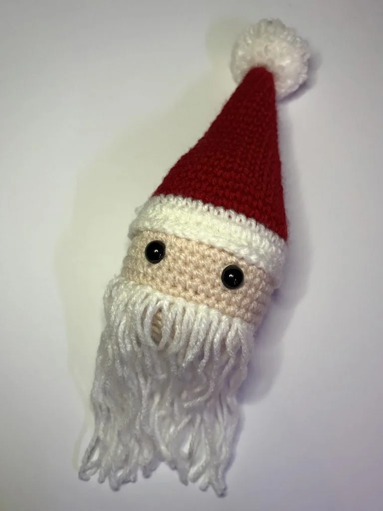 A crocheted Santa Claus head with a red hat, white trim, black button eyes, and a long white yarn beard sits on a plain background—perfect to display alongside your favorite crochet pumpkin pattern projects.