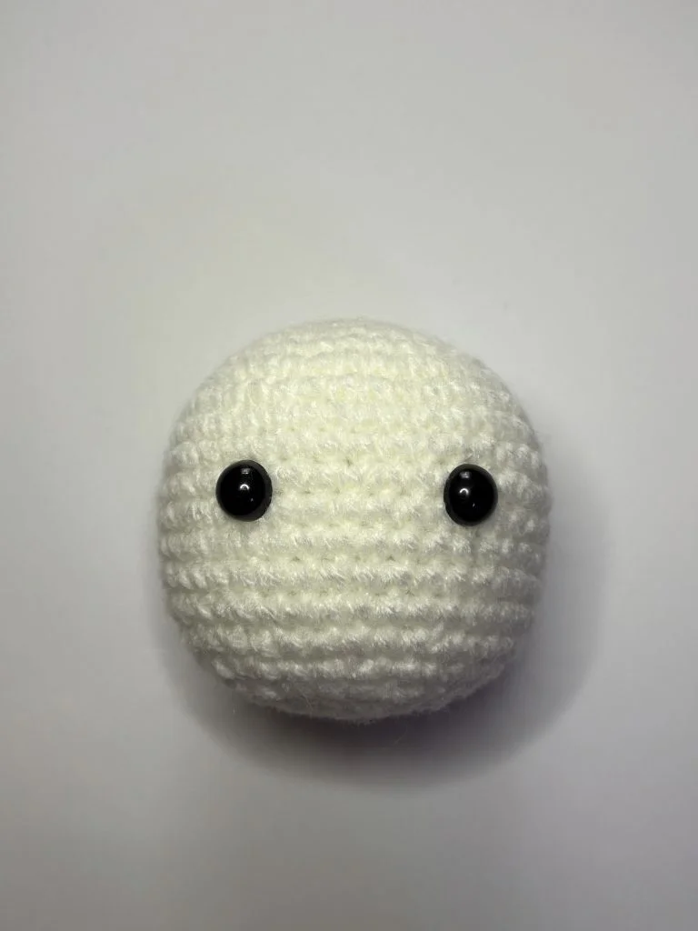A round, white crocheted ball with two black plastic eyes, placed on a plain white background, perfect for showcasing your latest pumpkin crochet pattern.