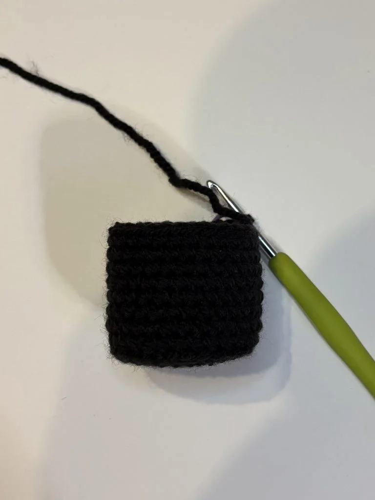 A small black crocheted square in progress with a green-handled crochet hook attached, resting on a white surface—perfect for starting your next pumpkin crochet pattern.