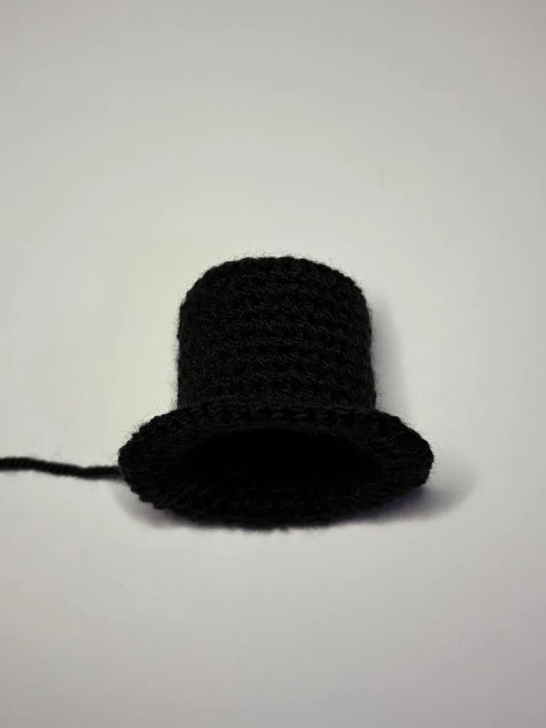 A small, black, crocheted top hat sits on a plain white surface with a strand of yarn attached—perfect for accenting your next crochet pumpkin pattern.