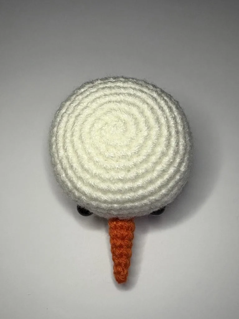 A crocheted white circle with an orange cone attached and two black beads, resembling a simplified snowman face, is displayed on a plain background—a whimsical touch to pair with your favorite pumpkin crochet pattern.
