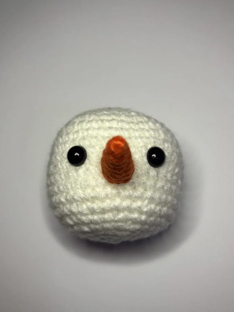 A crocheted snowman head with black button eyes and an orange carrot-shaped nose, photographed against a plain white background—perfect for makers who also enjoy crafting from a pumpkin crochet pattern.