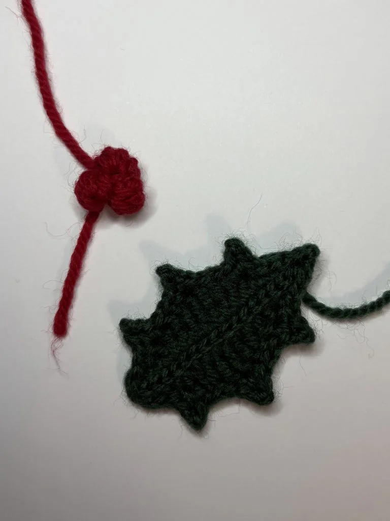 A crocheted dark green holly leaf and a small red berry are attached to red and green yarn, creating a festive piece that pairs beautifully with a crochet pumpkin or your favorite crochet pumpkin pattern, set against a white background.