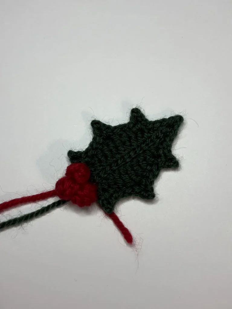 A crocheted green holly leaf with two red berries, set against a plain white background—perfect as a festive accent or to pair with your favorite crochet pumpkin pattern.