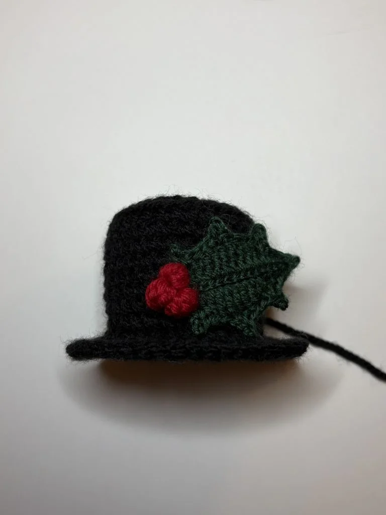 A small black crocheted hat with a green leaf and three red berries decoration attached to one side, photographed on a plain white background—perfect for pairing with your favorite pumpkin crochet pattern.