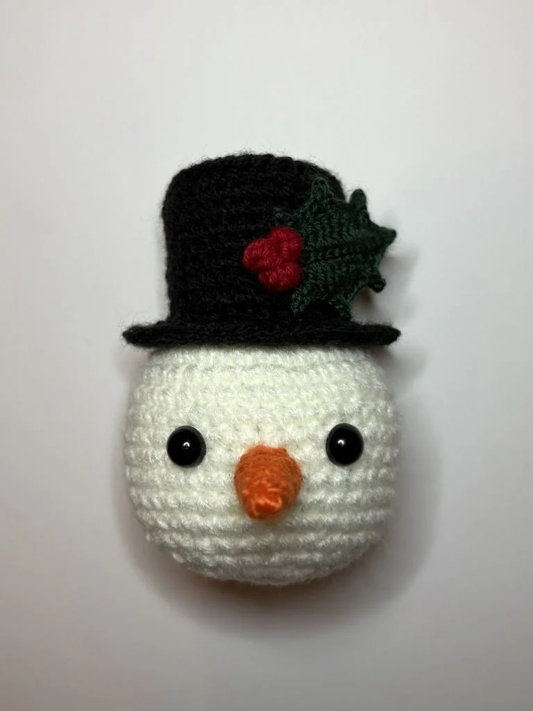 A crocheted snowman head with a black top hat, green holly leaf, red berries, black eyes, and an orange carrot nose—perfect alongside your crochet pumpkin creations—on a plain white background.