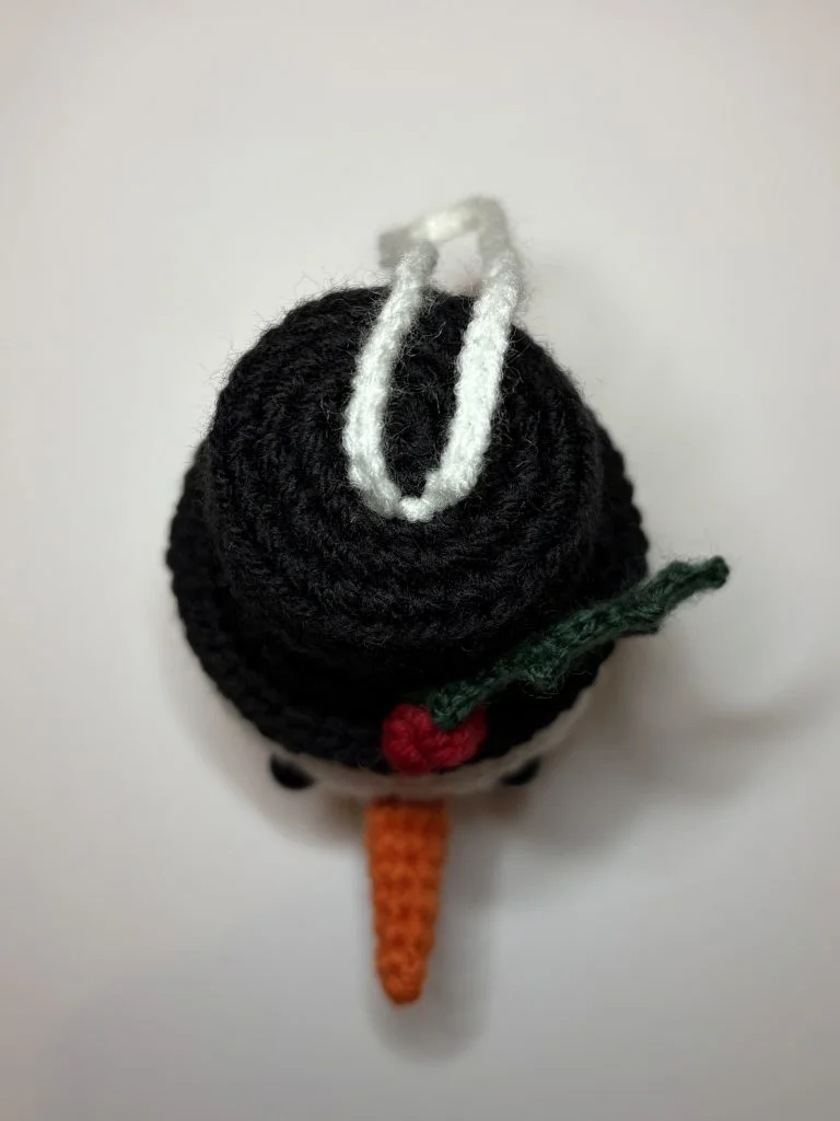A crocheted snowman ornament viewed from above, featuring a black hat with a white loop, a green leaf, red berry, an orange carrot nose—perfect alongside any crochet pumpkin or handmade holiday décor.