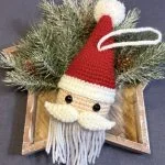 A crocheted Santa Claus ornament with a red hat, white beard, and mustache lies on pine branches in a wooden star-shaped tray—perfect for festive decor or as inspiration alongside your favorite free crochet pumpkin pattern.