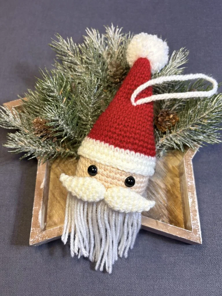 A crocheted Santa Claus ornament with a red hat, white beard, and mustache lies on pine branches in a wooden star-shaped tray—perfect for festive decor or as inspiration alongside your favorite free crochet pumpkin pattern.