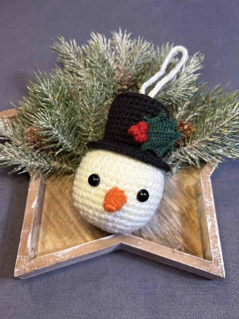 A crocheted snowman ornament with a black hat and holly decoration sits on a wooden star-shaped tray, surrounded by artificial pine branches, alongside a charming crochet pumpkin made using a free crochet pumpkin pattern.