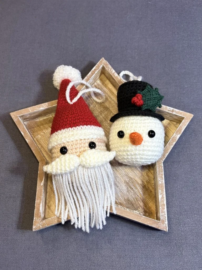 Crocheted Santa and snowman ornaments with button eyes rest on a wooden star-shaped tray against a gray background, perfect to display alongside your favorite crochet pumpkin pattern creations.