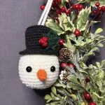 A crocheted snowman ornament with a black hat and orange nose hangs beside holly leaves, red berries, frosted pinecones, and would pair perfectly with a festive project made from a free crochet pumpkin pattern.