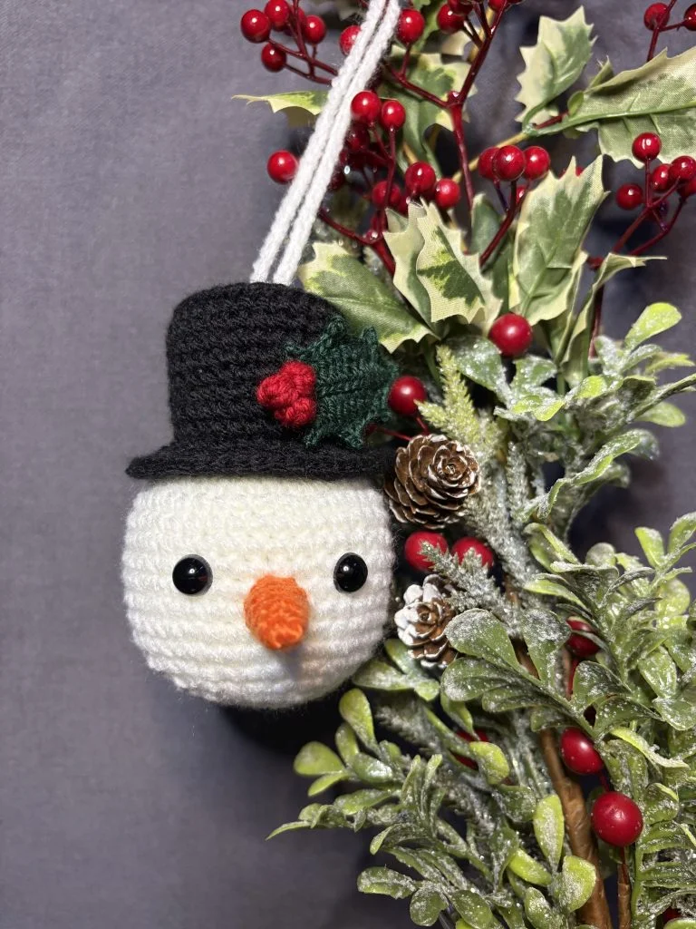A crocheted snowman ornament with a black hat and orange nose hangs beside holly leaves, red berries, frosted pinecones, and would pair perfectly with a festive project made from a free crochet pumpkin pattern.
