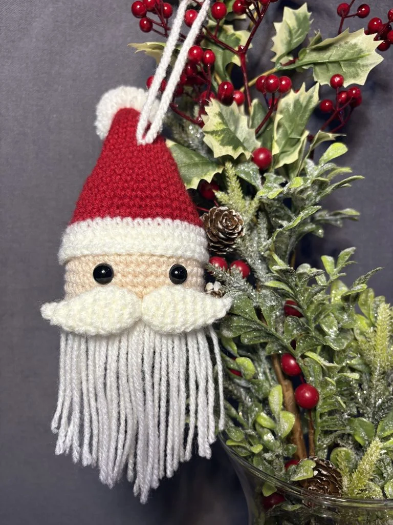 A crocheted Santa Claus ornament with a red hat, white mustache, and long yarn beard hangs beside festive greenery, red berries, and pinecones in a glass vase—a cozy touch for fans of any crochet pumpkin pattern.