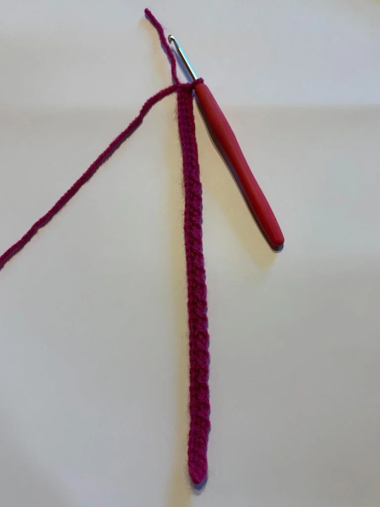 A crochet hook with a red handle is attached to a length of magenta yarn and a completed foundation chain—perfect for starting your next pair of crochet fingerless gloves—on a light surface.