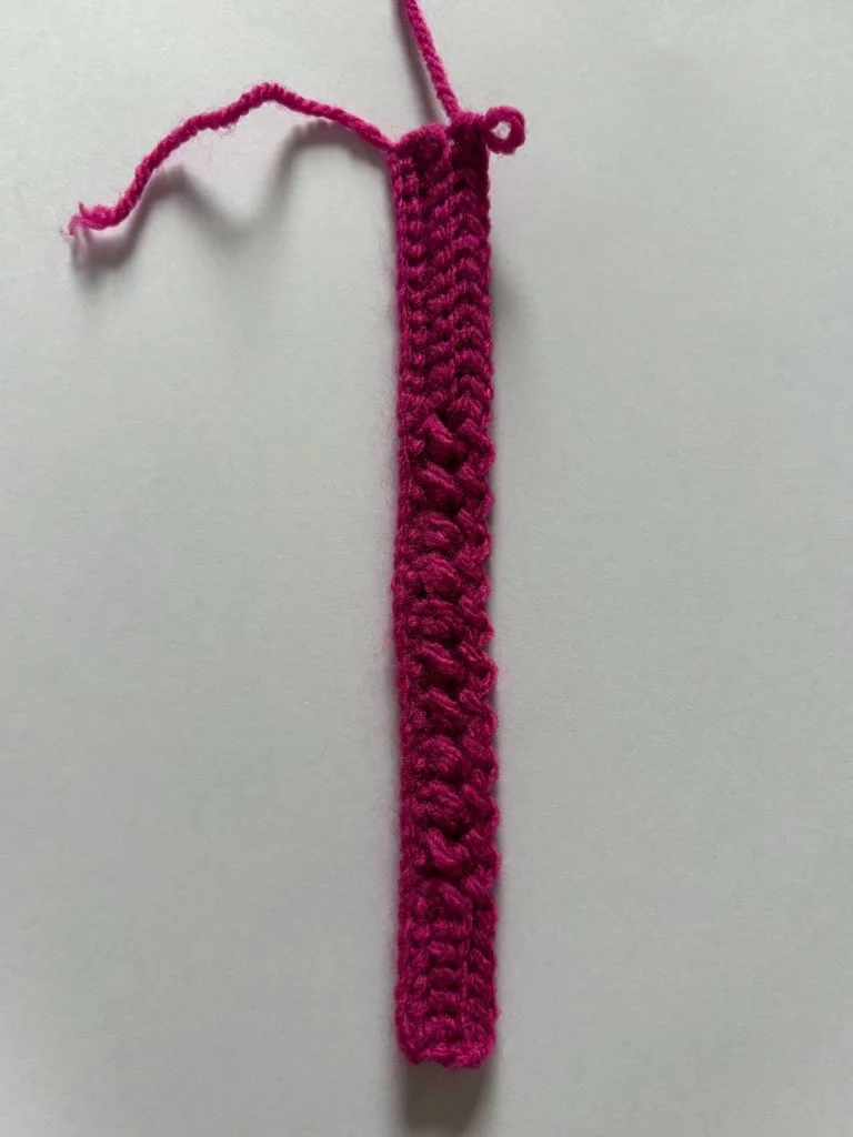 A narrow strip of bright pink crochet, possibly the start of crochet fingerless gloves, with a loose yarn end is displayed on a plain white background.