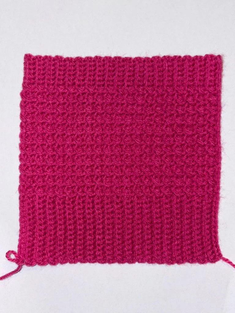 A square swatch of textured pink knitting with a ribbed bottom edge and a patterned upper section, perfect inspiration for crochet fingerless gloves, displayed on a white background.