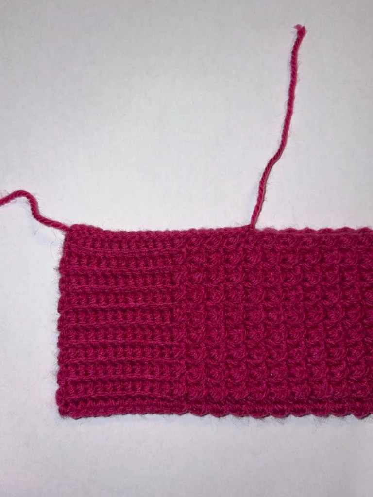 A rectangular piece of pink crochet, ideal for making crochet fingerless gloves, features two different stitch patterns and two yarn tails on a white surface.