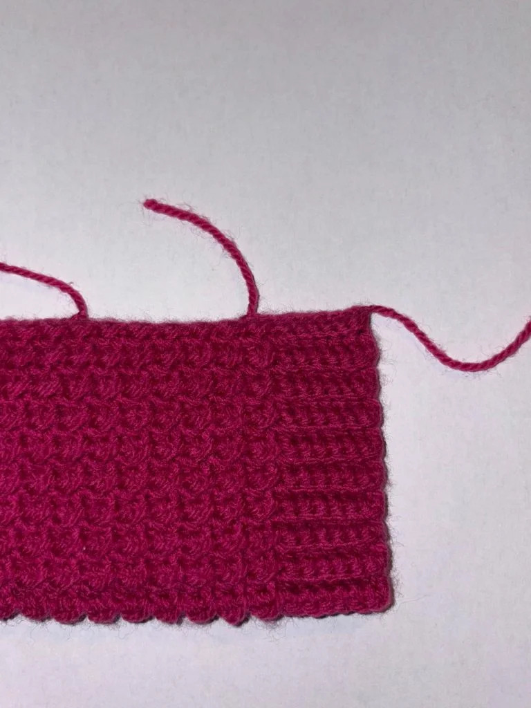 A piece of textured pink knitting, possibly destined to become crochet fingerless gloves, with two loose yarn ends on a plain white background.