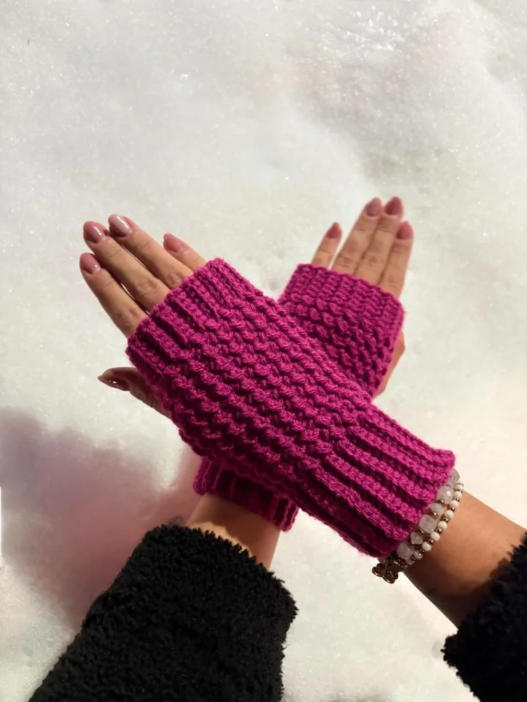Hands wearing textured, magenta crochet fingerless gloves are crossed over each other on a snowy background. The person also wears a bracelet with white beads and a black sleeve.