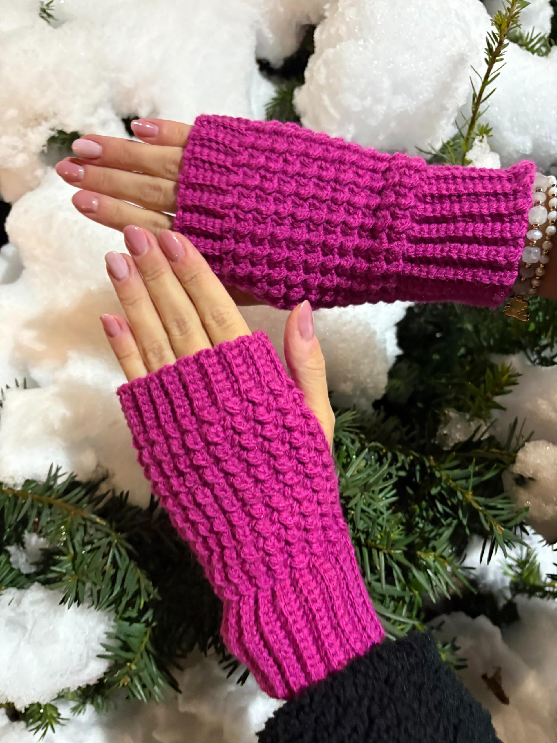 Two hands wearing crochet fingerless gloves in magenta are held over snow-covered greenery.