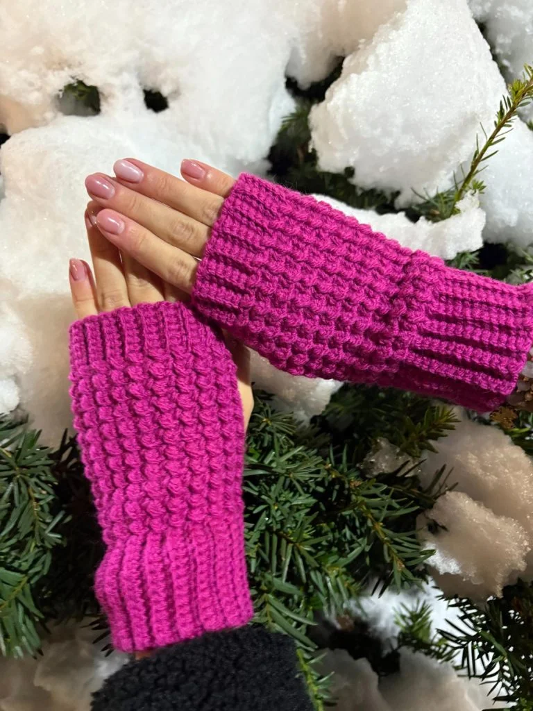 Hands wearing bright pink crochet fingerless gloves are held over snow-covered green foliage.