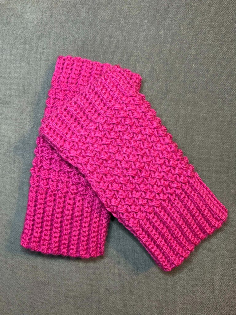 A pair of bright pink, hand-knitted crochet fingerless gloves with a textured pattern, laid out on a gray surface.