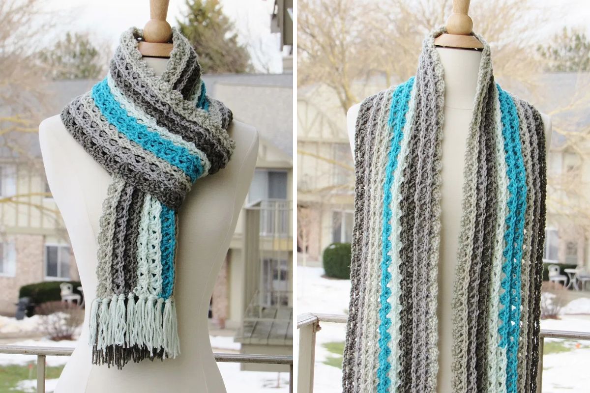 The Ocean Waves Scarf, with its gray, white, and blue stripes, is draped around a dress form. Braided fringe accents one end, while snow and trees create a wintry backdrop outside.