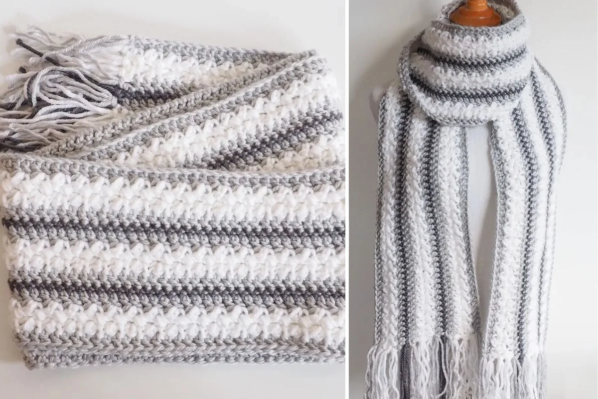 A crocheted scarf with white, light gray, and dark gray horizontal stripes, featuring a textured pattern and fringe at the ends.