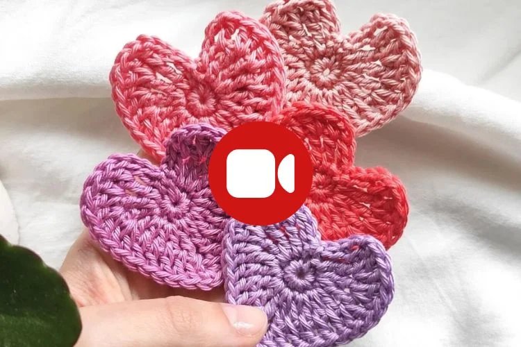 A hand holds five crocheted heart shapes in pink, red, and purple tones. A red video call icon is centered over the hearts.