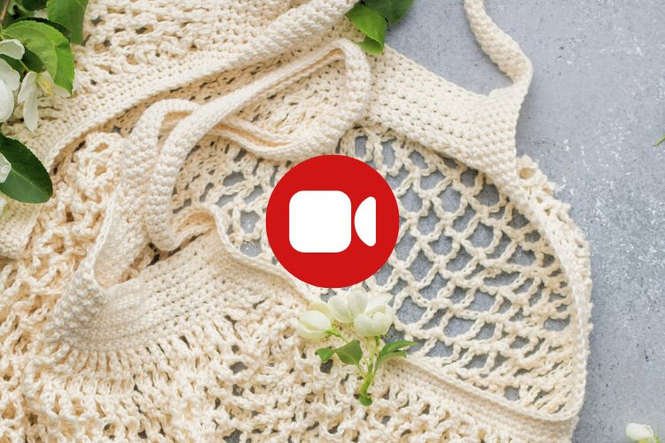 A cream-colored crocheted bag with green leaves and white flowers on a gray surface; a red video camera icon is centered on the image.