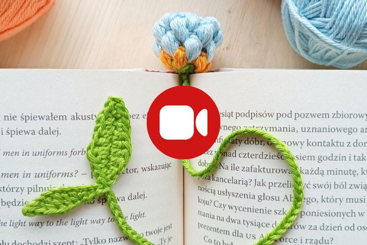 An open book with a crocheted flower and leaf bookmark placed across its pages; balls of yarn are visible in the background.